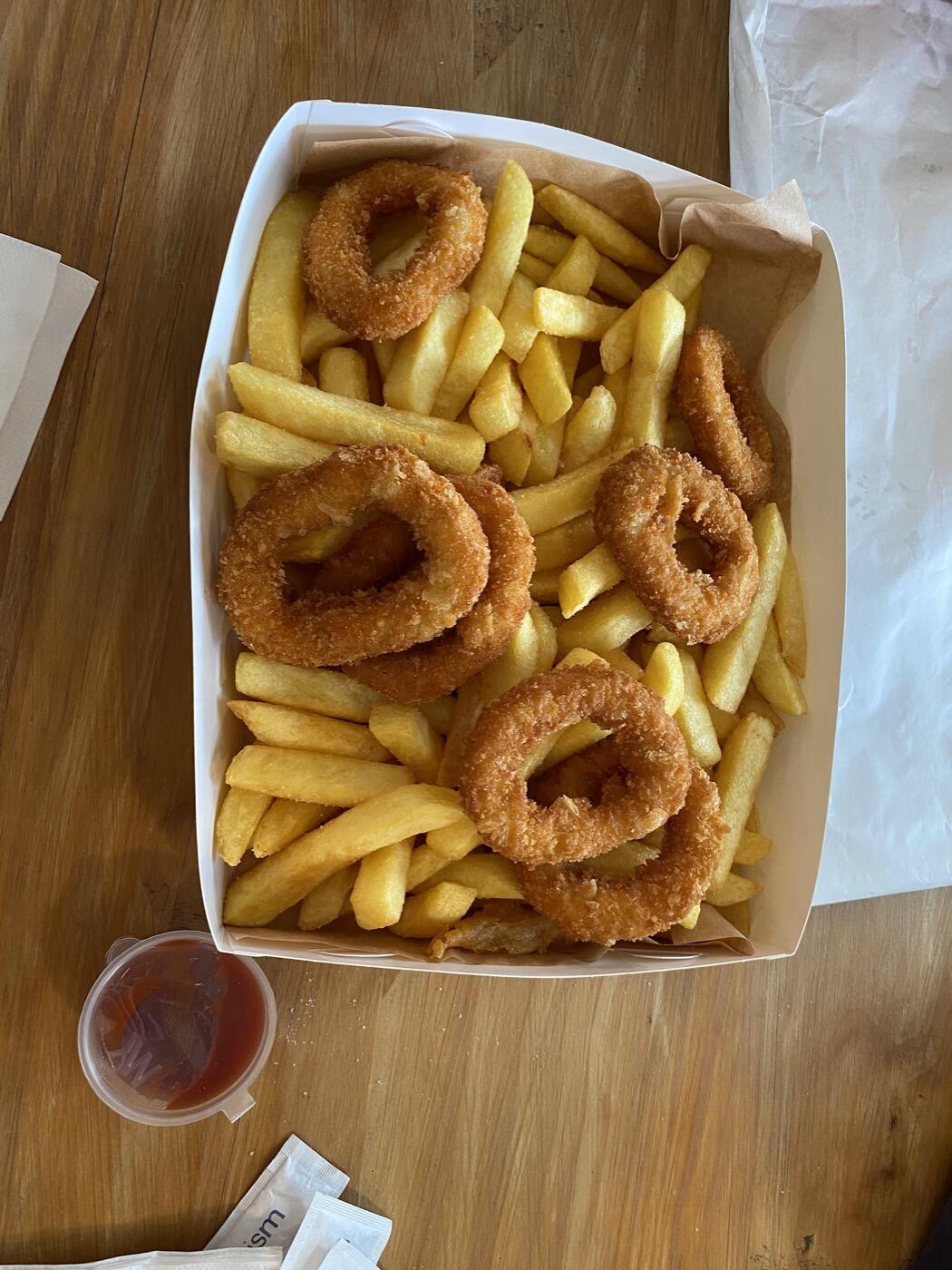 A cardboard box of chips and crumbed calamari rings on a timber table, with a small cup of red sauce