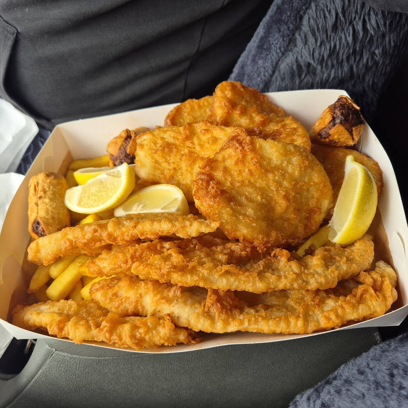 A cardboard tray piled with golden battered fish fillets and chips, with lemon wedges tucked in at the side