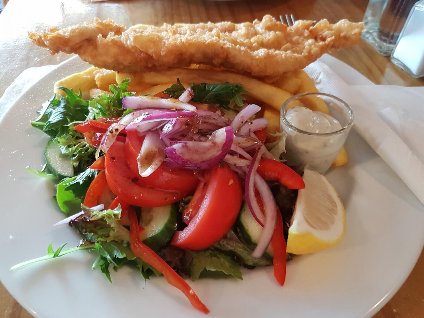 A large piece of battered fish over chips on a white plate, with a salad of tomato, cucumber and red onion, a lemon wedge and a pot of sauce