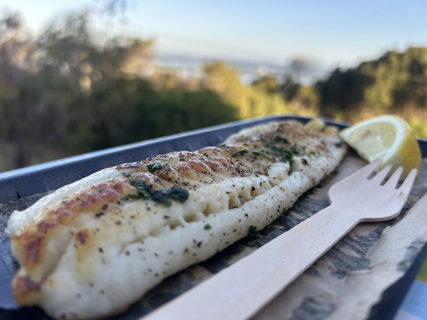 A grilled white fish fillet with herbs and a lemon wedge on a tray with a wooden fork, with trees and the bay blurred behind