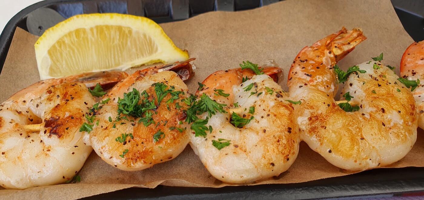 A row of grilled prawns scattered with chopped parsley on baking paper, with a lemon wedge