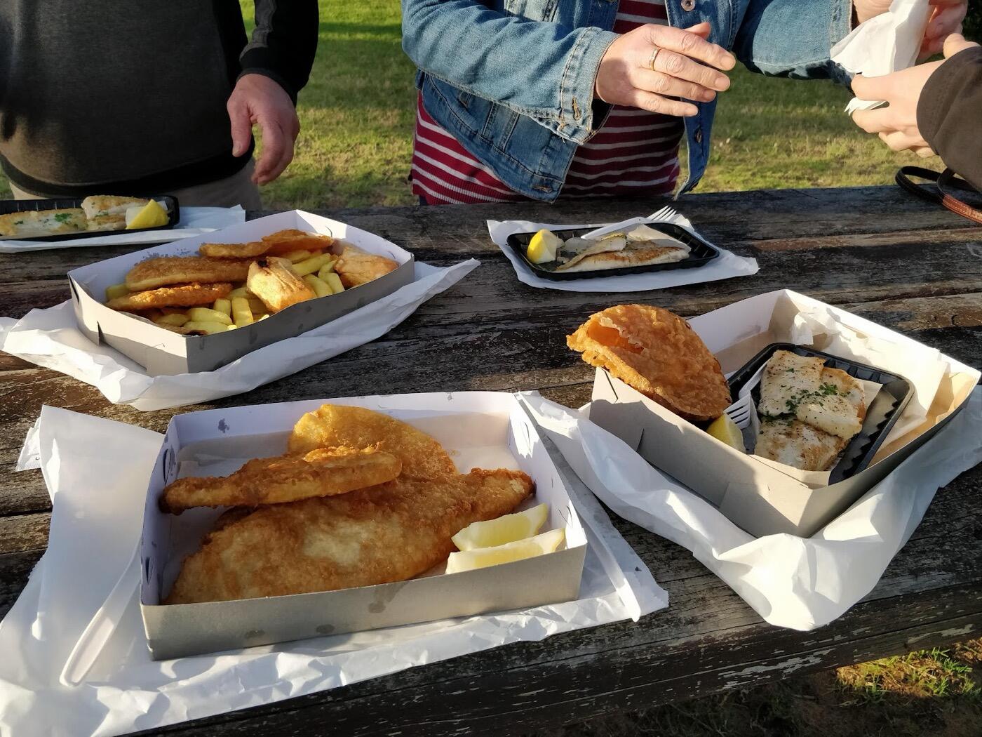 Cardboard boxes of battered fish and chips and a tray of grilled fish on a wooden picnic table outdoors
