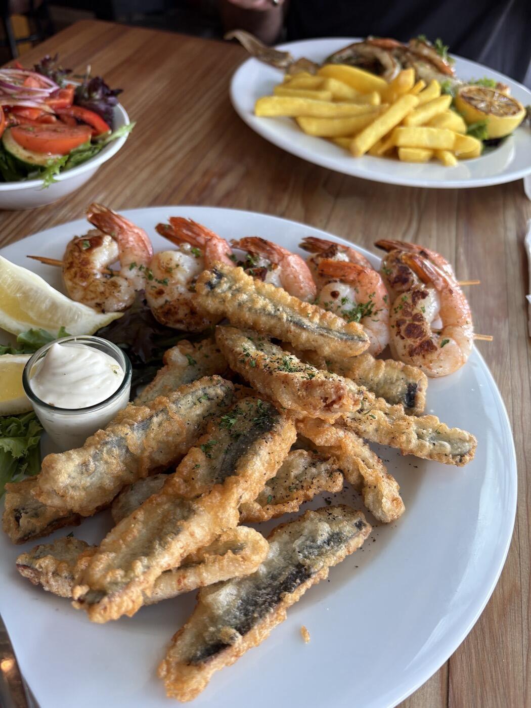 A white plate of fried sardines and prawn skewers with a small pot of white dipping sauce, with a salad and chips on the table behind